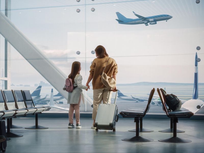 Mulher e criança de mãos dadas de costas e segurando uma mala de viagem, olhando um avião que decola ao fundo